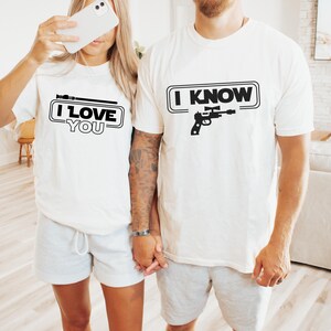 May include: A couple wearing matching white t-shirts with a Star Wars themed graphic. The woman's shirt says "I Love You" with a lightsaber graphic. The man's shirt says "I Know" with a blaster graphic.