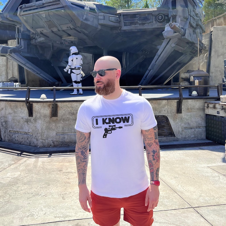 May include: A white t-shirt with a black graphic of a blaster and the text "I KNOW". The person wearing the shirt has a beard and is standing in front of a large, gray spaceship.