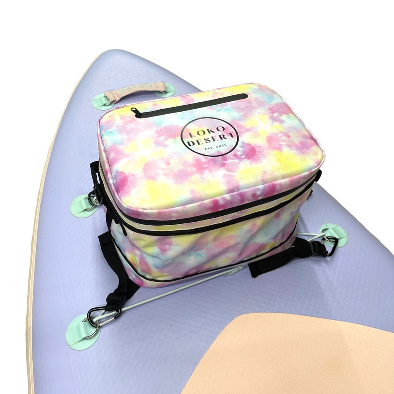 May include: A tie-dye cooler bag with black straps and a black zipper. The bag has a white circle logo with the text "LOKO DESERT EST. 2021". The bag is attached to a light purple and beige inflatable paddleboard.
