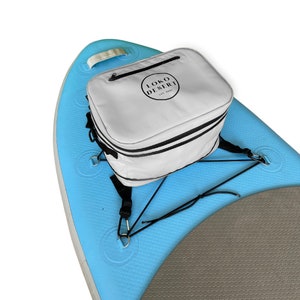 May include: A white cooler bag with black straps and the text "LOKO DESERT EST. 2022" attached to a blue and gray inflatable paddleboard.