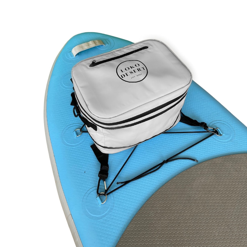 May include: A white cooler bag with black straps and the text "LOKO DESERT EST. 2022" attached to a blue and gray inflatable paddleboard.