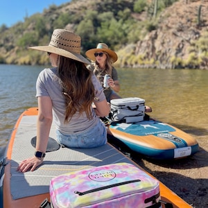 May include: Two people on paddleboards on a lake. One person is wearing a straw hat and the other is wearing a green shirt. There is a tie-dye cooler bag with the text "LOKO DISRUPT" on it.