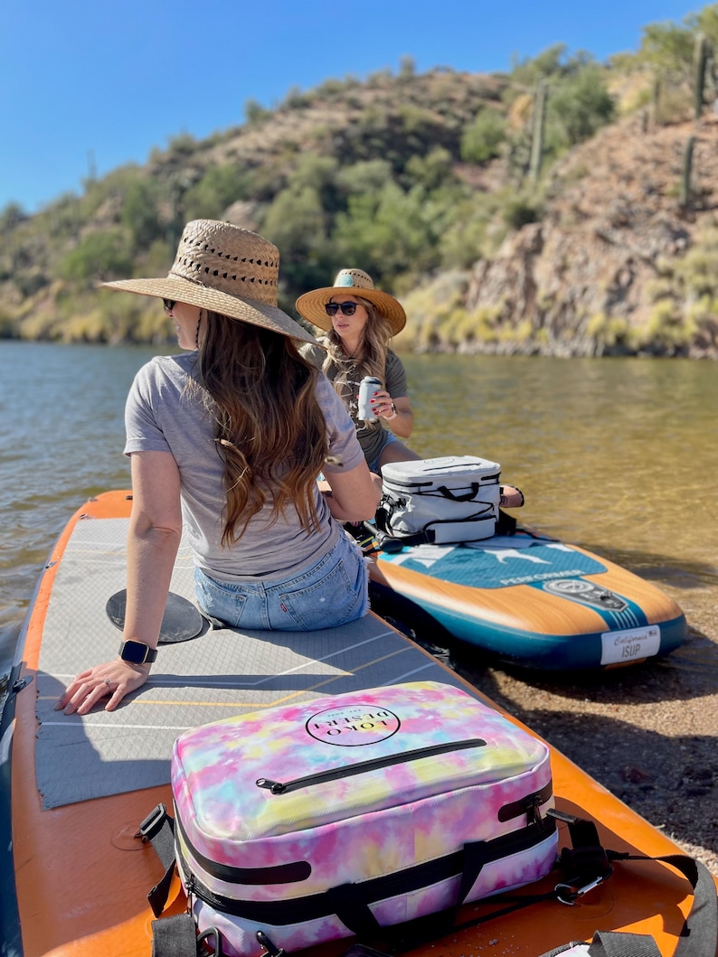 May include: Two people on paddleboards on a lake. One person is wearing a straw hat and the other is wearing a green shirt. There is a tie-dye cooler bag with the text "LOKO DISRUPT" on it.
