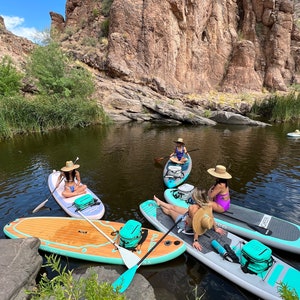 May include: Four people on turquoise and white stand up paddle boards in a calm lake with a rocky cliff in the background. The paddle boards have a turquoise stripe and a black and white logo. Each paddle board has a cooler bag.