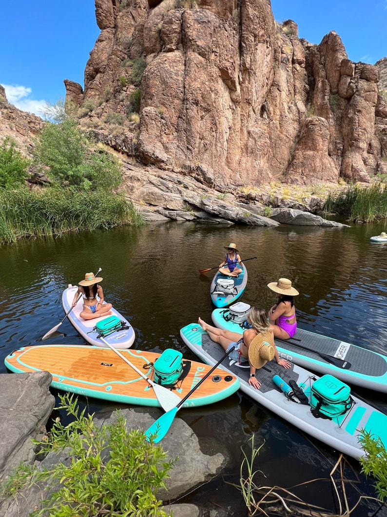 May include: Four people on turquoise and white stand up paddle boards in a calm lake with a rocky cliff in the background. The paddle boards have a turquoise stripe and a black and white logo. Each paddle board has a cooler bag.