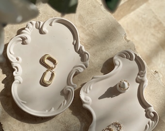 May include: Two off-white, ornate, scalloped-edge trays hold gold jewellery. One tray displays a pair of rectangular gold hoop earrings. The other tray holds a small white ring stand with a gold ring and two additional gold rings nearby. Text reads CandlesCo.
