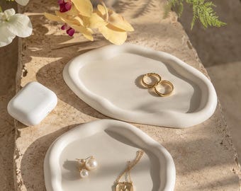 May include: Two white, organic-shaped, matte-finish trays hold jewelry. The top tray contains two gold rings. The bottom tray holds a pair of pearl stud earrings and a gold pendant necklace. A white wireless earbud case sits to the left.