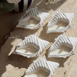 May include: Five white ceramic seashell candle holders with a white candle in each. The candles are on a light brown surface. The text "CandlesCo" is visible in the bottom right corner of the image.
