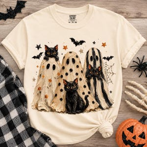 May include: A cream-colored short-sleeve t-shirt featuring a graphic print of three black cats wearing ghost costumes. The design includes small orange flowers, black polka dots, black stripes, bats, stars, and a spider web. The shirt has a Comfort Colors label.