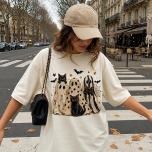 May include: A beige baseball cap and an oversized cream t-shirt featuring a graphic print of three black cats dressed as ghosts, surrounded by small orange stars, black bats, and a spider web design, paired with a black shoulder bag with a chain strap.