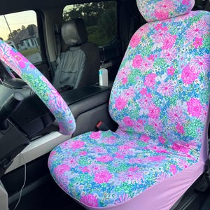 May include: Floral car seat cover and headrest cover with a matching steering wheel cover. The seat cover features a vibrant pattern of pink and blue flowers on a white background. The steering wheel cover has a similar design.