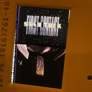 May include: A yellow plastic 35mm film slide mount featuring a Star Trek First Contact movie title graphic against a starry space background. The frame includes printed text reading NCN 855X00075LOT75 and 10/96 (816)761-4001 along the left side.
