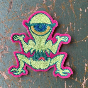 Op de afbeelding: Geborduurde patch met een cartoon alien met één oog. De alien is limoengroen met een blauw en geel oog, omlijnd door een felroze rand. De patch heeft een grillig ontwerp, geschikt voor kleding of accessoires.