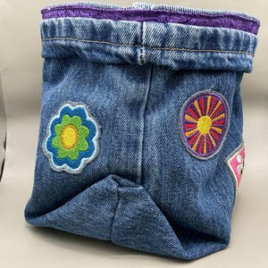 May include: A blue denim fabric storage bin featuring a folded top edge with a purple patterned interior lining. The exterior is decorated with embroidered patches, including a green and yellow flower and a circular sunburst design in orange and purple.