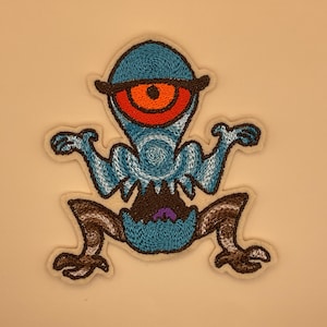 May include: Embroidered patch featuring a cartoonish, one-eyed creature in shades of blue, brown, and orange. The creature has a large eye, a wide mouth, and outstretched arms and legs. The patch is outlined with a cream-colored border.