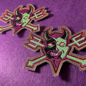 Może przedstawiać: Dwie haftowane naszywki z motywem diabła. Diabeł ma podzieloną twarz, z jedną stroną czarną, a drugą zieloną. Naszywki mają fioletowe tło, zielone i pomarańczowe akcenty oraz szarą obwódkę.