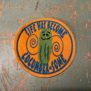 May include: An orange circular patch with blue embroidered text: "LIFE HAS BECOME CUCUMBER-SOME." A green pickle with a smiling face is in the center. The patch has a felt-like texture.