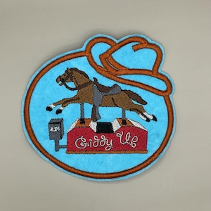 Op de afbeelding: Geborduurde patch met een bruin paard op een rood en zwart platform met de tekst "Giddy Up". De patch heeft een lichtblauwe achtergrond met een bruine rand en een bruin touwontwerp. Een klein vakje leest "25¢".