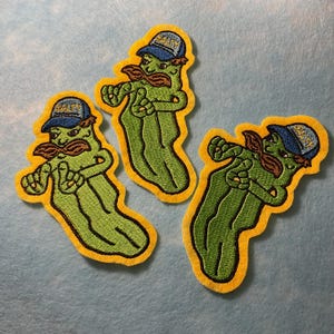 May include: Three embroidered patches of a cartoon pickle character. The pickle is green with a brown moustache and a blue cap with the word "Salty". Each patch has a yellow border.