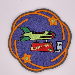 Könnte beinhalten: Gestickter Aufnäher mit einem Cartoon-Raumschiff und dem Text "BLAST OFF!" in Weiß. Der Aufnäher hat eine lila Mitte, einen orangefarbenen Rand und zwei orangefarbene Sterne. Das Design ist kreisförmig.