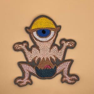 Peut inclure: Écusson brodé représentant une créature fantaisiste à un œil, coiffée d'un chapeau jaune, avec un œil bleu et les bras tendus. Le patch a une bordure grise et est réalisé avec des fils roses, bruns et bleus. Le design est sur fond beige.