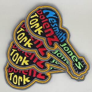 May include: Embroidered patches in a guitar shape, stacked on top of each other. The patches have a black background with colorful text that reads "Dolez Tork" and "Yesmith Jones". The patches have a gray border with a gold outline.