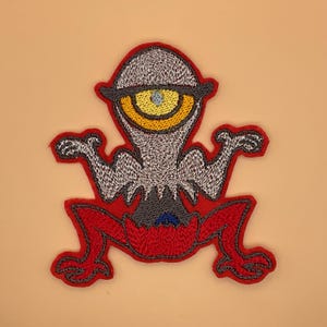 May include: Embroidered patch featuring a cartoon alien with a large, yellow and blue eye. The alien has a gray head and body, red legs, and is outlined in red. The patch is designed for application on clothing or accessories.