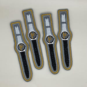 Könnte beinhalten: Vier gestickte Aufnäher in Form einer Uhr. Jeder Aufnäher hat ein graues Zifferblatt mit einem schwarzen Armband, einem weißen Band und goldenen Akzenten. Das Zifferblatt hat ein goldenes Design. Die Aufnäher sind gold- und grau umrandet.