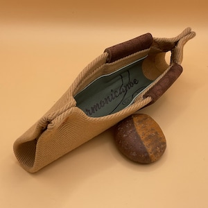 Pode incluir: Um estojo de harmónica em tecido bege com detalhes em couro castanho e uma pequena pedra arredondada. O estojo tem uma textura tecida e o texto "HarmonicaCanoe" é visível no interior. A pedra é castanha e bege.