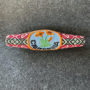 May include: A woven hair barrette with a California patch. The patch features orange flowers and the word "California" in white lettering on a blue background. The barrette has a red, pink, and black geometric pattern.