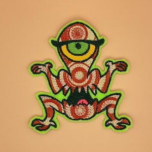 May include: Embroidered patch featuring a whimsical, one-eyed creature with a red and white striped body, a green eye, and a black brow. The patch has a neon green border and an open mouth with a pink tongue. The design is on a tan background.