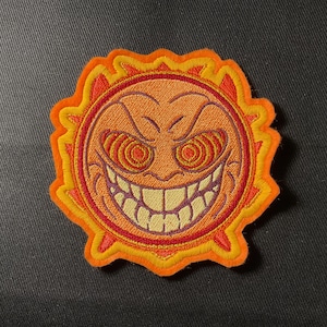 Op de afbeelding: Geborduurde patch met een gestileerde zon met een boze uitdrukking. De zon heeft een feloranje gezicht met wervelende ogen, een brede grijns en een gekartelde, gele omtrek. De patch is omrand door een rood en geel ontwerp.
