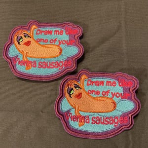 Puede incluir: Dos parches bordados con un diseño de salchicha vienesa de dibujos animados. La salchicha tiene una cara y está sobre una nube azul. El texto dice "Draw me like one of your Vienna sausages" en rosa.