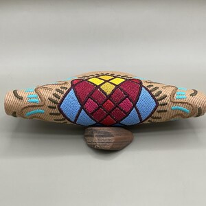 May include: A tan, oblong decorative object with detailed embroidery. The central design features a geometric pattern in red, blue, and yellow. The ends have turquoise and brown embroidered patterns. It rests on a small, brown, stone-like base.