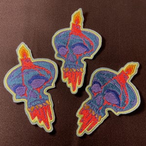 Puede incluir: Tres parches bordados con un diseño de calavera con una vela. Las calaveras son azules y moradas con cera roja goteando. Los parches tienen un borde amarillo claro.