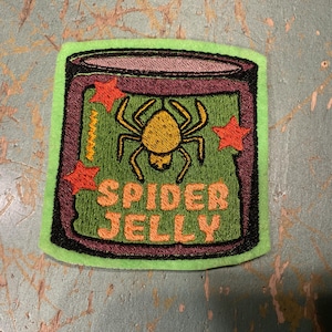 Puede incluir: Parche bordado con borde verde. El diseño presenta un tarro con una araña amarilla y las palabras "SPIDER JELLY" en naranja. Estrellas rojas adornan el tarro, sobre un fondo verde.