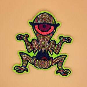 Puede incluir: Parche bordado con una criatura colorida de un ojo con un contorno verde neón. La criatura tiene un ojo grande con un patrón en espiral, y su cuerpo está lleno de hilos arremolinados de varios colores. El parche está sobre un fondo beige.