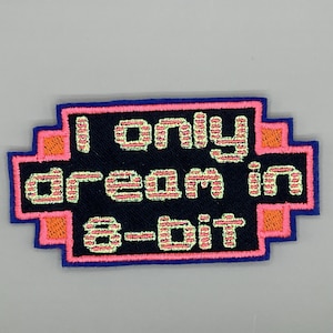 May include: Embroidered patch with the text "I only dream in 8-bit." The patch has a black background with pink and orange accents. The border is blue.