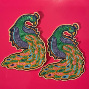 Könnte beinhalten: Zwei gestickte Pfauen-Patches. Jeder Patch hat einen grünen Kopf und Hals, einen blauen und lila Körper und einen fächerförmigen Schwanz mit rosa, blauen und orangefarbenen Details. Die Patches haben einen cremefarbenen Rand und sind vor einem hellrosa Hintergrund platziert.