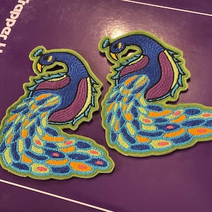 May include: Two embroidered peacock patches. Each patch features a blue head and neck, a purple chest, and a vibrant tail with blue, yellow, orange, and pink details. The patches are outlined in green.