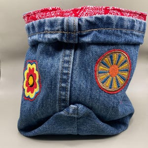 May include: A blue denim fabric storage basket featuring a folded top edge lined with red and white paisley patterned fabric. The exterior is decorated with two embroidered patches: a yellow and red flower and a circular sunburst design.