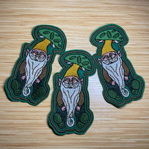 May include: Three embroidered patches featuring a gnome with a white beard, wearing a yellow hat and brown coat, under a green mushroom cap. The patches have a green border and are on a light brown surface.