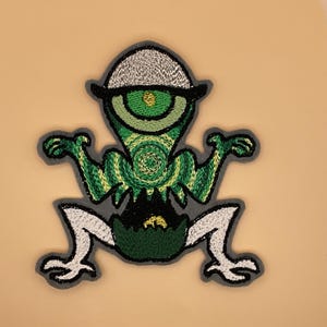 Peut inclure: Écusson brodé représentant une créature verte à un œil, coiffée d'un chapeau. Le motif utilise des fils verts, jaunes, noirs et blancs, avec une bordure grise. La créature a les bras et les jambes tendus, ce qui lui donne un aspect ludique.