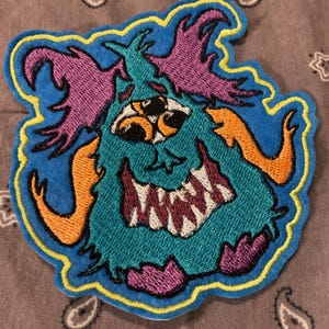 Puede incluir: Parche bordado con un monstruo de dibujos animados con pelaje turquesa, cuernos naranjas y detalles morados. El monstruo tiene tres ojos y una amplia sonrisa con dientes. El parche está delineado en amarillo y azul.
