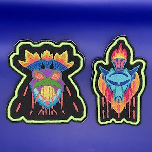 Puede incluir: Dos parches bordados con bordes verde neón. Uno presenta una cara de monstruo colorida con una boca ancha y dientes grandes. El otro parche representa una criatura azul y naranja con llamas. Ambos parches están sobre un fondo oscuro.