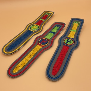 May include: Three embroidered watch patches in blue, green, and yellow. Each patch features a watch face with a colorful design and a matching strap. The patches are outlined in gray and have a yellow border.