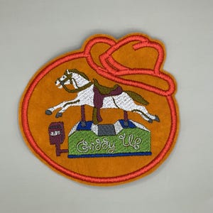 May include: Embroidered patch with a carousel horse in white and brown, with a saddle and reins, set against an orange background. The patch includes a red border and a cowboy hat design.