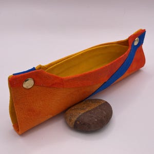 May include: A vibrant, handmade fabric pencil case with orange, yellow, and blue details. The case is fastened with gold-coloured press studs. A small, two-toned stone sits nearby.