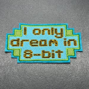 May include: A rectangular patch with a pixelated design and the text "I only dream in 8-bit." The patch has a light blue background with green and yellow accents, and the text is in yellow.