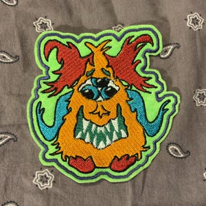 Könnte beinhalten: Gestickter Aufnäher mit einem Cartoon-Monster mit drei Augen, orangefarbenem Fell, roten Hörnern und grünen Konturen. Das Monster hat ein breites, zahnreiches Grinsen und blaue Akzente. Der Aufnäher befindet sich auf einem grauen Paisley-Hintergrund.